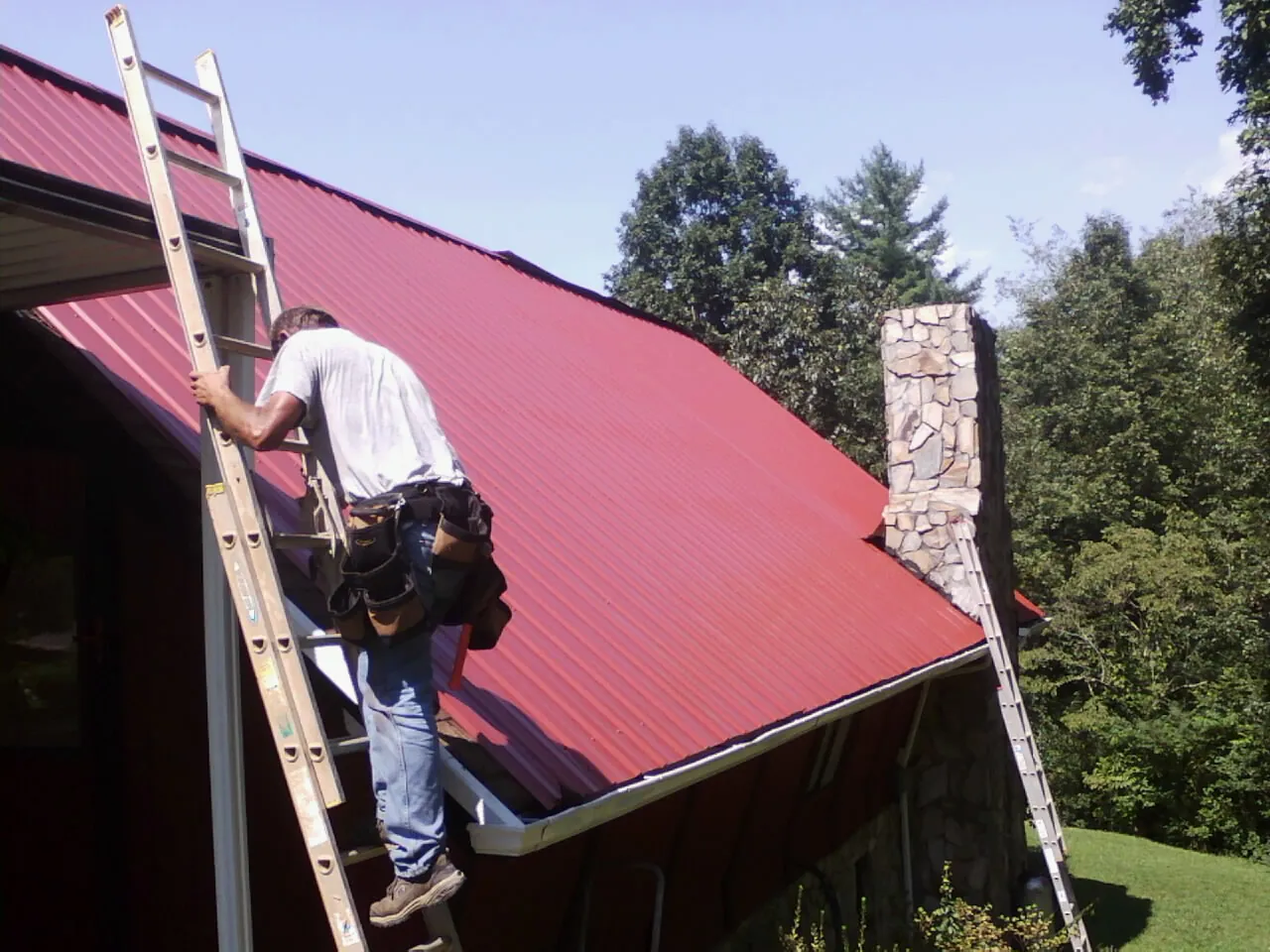 Roofer on a ladder beside a red metal roof and stone chimney — Soffit & Fascia Repair by Roofing Brothers in Landisville